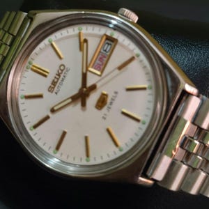 Puede incluir: Un reloj de pulsera autom&aacute;tico Seiko plateado con esfera blanca, manecillas e indicadores dorados, y visualizaci&oacute;n de d&iacute;a y fecha. El reloj tiene una correa plateada y muestra el domingo 8. La esfera dice "SEIKO AUTOMATIC 21 JEWELS".
