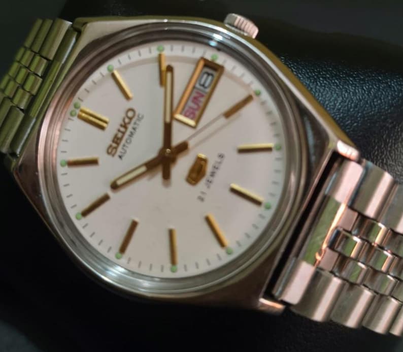 Puede incluir: Un reloj de pulsera autom&aacute;tico Seiko plateado con esfera blanca, manecillas e indicadores dorados, y visualizaci&oacute;n de d&iacute;a y fecha. El reloj tiene una correa plateada y muestra el domingo 8. La esfera dice "SEIKO AUTOMATIC 21 JEWELS".