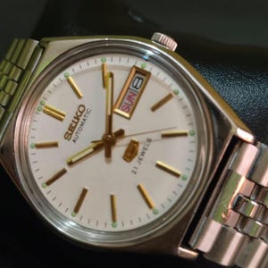 Puede incluir: Un reloj de pulsera autom&aacute;tico Seiko vintage con caja y brazalete plateados. La esfera es blanca con marcadores y agujas doradas. El d&iacute;a y la fecha se muestran a las 3 en punto. El reloj est&aacute; marcado "SEIKO AUTOMATIC 21 JEWELS".