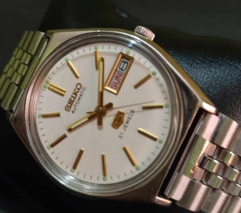 Puede incluir: Un reloj de pulsera autom&aacute;tico Seiko vintage con caja y brazalete plateados. La esfera es blanca con marcadores y agujas doradas. El d&iacute;a y la fecha se muestran a las 3 en punto. El reloj est&aacute; marcado "SEIKO AUTOMATIC 21 JEWELS".