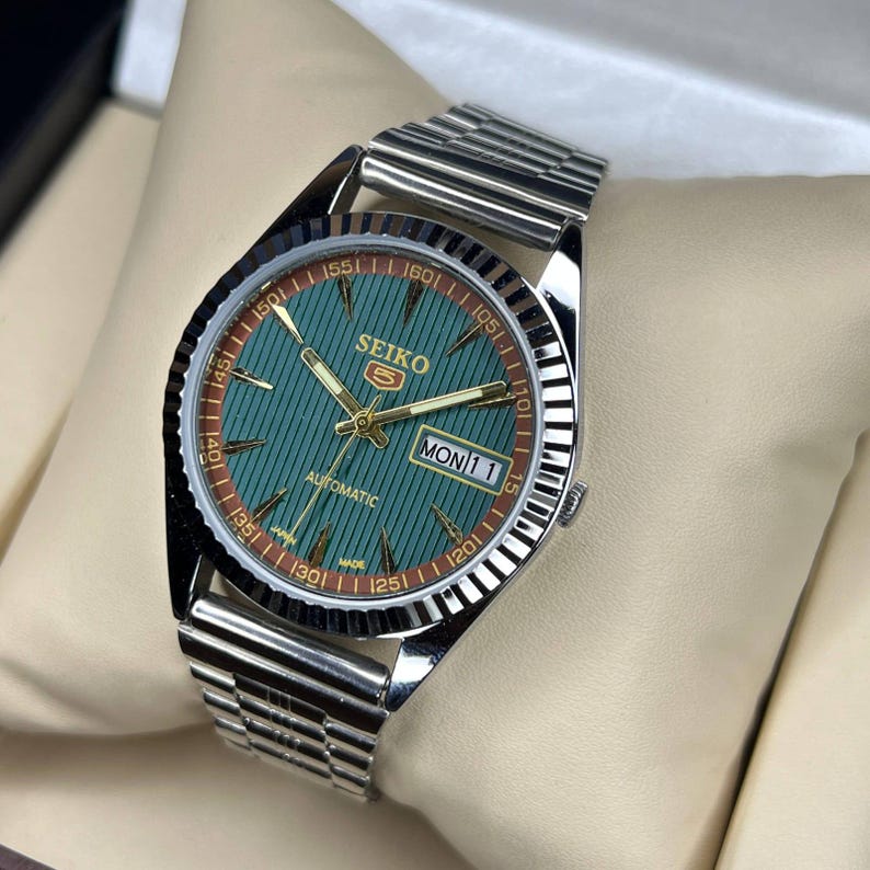 May include: A silver-tone Seiko 5 automatic watch with a textured teal green dial, gold-tone hands and indices, and a fluted bezel. The watch features a day-date window at the three o'clock position and a silver-tone metal link bracelet strap.