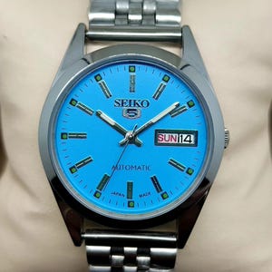 May include: A silver-tone stainless steel Seiko 5 automatic wristwatch featuring a bright blue dial with luminous silver-tone hour markers and hands. The watch includes a day-date window at the three o'clock position and a metal link bracelet.