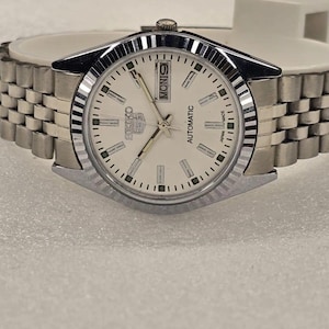 May include: A silver-tone Seiko automatic wristwatch with a white dial, silver baton indices, and a fluted bezel. The watch features a day-date window at the three o'clock position, silver hands, and a multi-link metal bracelet with a fold-over clasp.