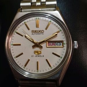 Puede incluir: Un reloj de pulsera autom&aacute;tico Seiko plateado con esfera blanca. El reloj presenta agujas e indicadores dorados, una pantalla de d&iacute;a y fecha y el texto "21 Jewels". La correa del reloj est&aacute; hecha de eslabones de metal plateado.