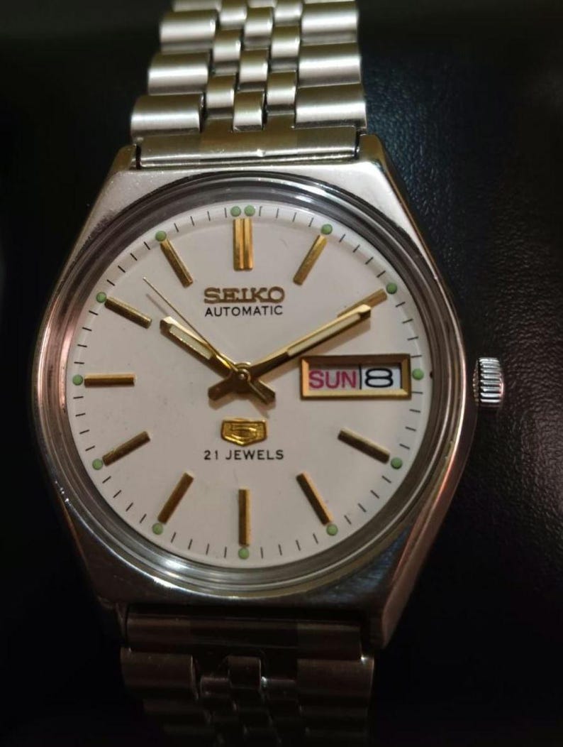 Puede incluir: Un reloj de pulsera autom&aacute;tico Seiko plateado con esfera blanca. El reloj presenta agujas e indicadores dorados, una pantalla de d&iacute;a y fecha y el texto "21 Jewels". La correa del reloj est&aacute; hecha de eslabones de metal plateado.