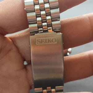 May include: Close-up of a stainless steel watch band with a Seiko clasp. The band features a repeating pattern of small, rectangular links. The Seiko logo is prominently displayed on the clasp in a rectangular shape.