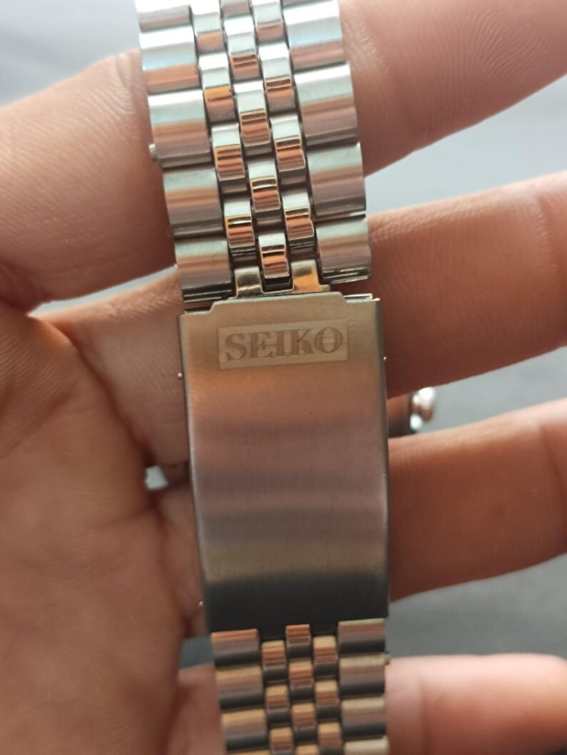 May include: Close-up of a stainless steel watch band with a Seiko clasp. The band features a repeating pattern of small, rectangular links. The Seiko logo is prominently displayed on the clasp in a rectangular shape.