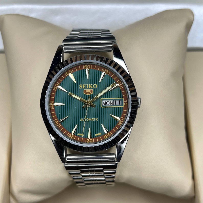 May include: A Seiko 5 automatic wristwatch featuring a silver-tone stainless steel link bracelet and a round case. The dial is green with vertical pinstripes, gold-tone hands and indices, and a day-date window at the three o'clock position.