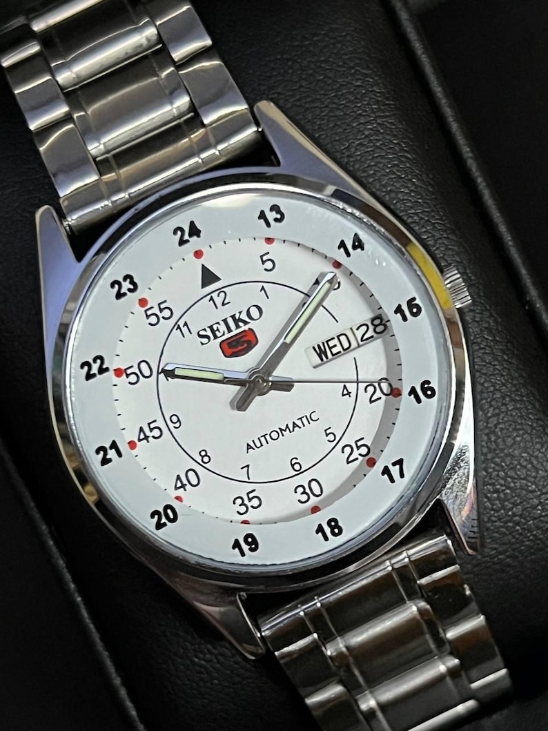 May include: A silver-toned Seiko automatic wristwatch with a white face. The watch features black numerals, a day-date display, and a silver band. The watch face displays the brand name and the word "AUTOMATIC".