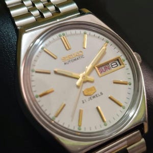 Puede incluir: Reloj de pulsera autom&aacute;tico Seiko plateado con esfera blanca, manecillas e indicadores dorados y visualizaci&oacute;n de d&iacute;a y fecha. El reloj tiene una correa de acero inoxidable y el texto "SEIKO AUTOMATIC" y "21 JEWELS".