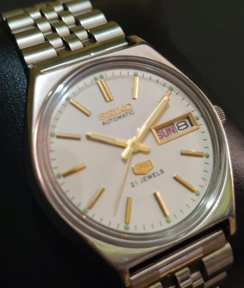 Puede incluir: Reloj de pulsera autom&aacute;tico Seiko plateado con esfera blanca, manecillas e indicadores dorados y visualizaci&oacute;n de d&iacute;a y fecha. El reloj tiene una correa de acero inoxidable y el texto "SEIKO AUTOMATIC" y "21 JEWELS".