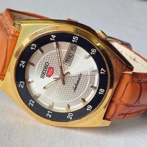 May include: A gold-tone Seiko 5 automatic wristwatch featuring a textured silver dial, a black outer bezel with 24-hour markings, and a brown leather strap with a crocodile-embossed pattern. The dial displays the Seiko logo, day, and date.