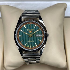 May include: A Seiko 5 automatic wristwatch featuring a silver-tone stainless steel link bracelet and a round case. The dial is green with vertical pinstripes, gold-tone hands and indices, and a day-date window at the three o'clock position.