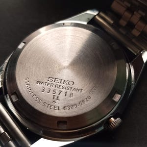 May include: Close-up of the back of a Seiko stainless steel watch. The watch face is silver with the words "SEIKO WATER RESISTANT" and other text. The watch band is silver.