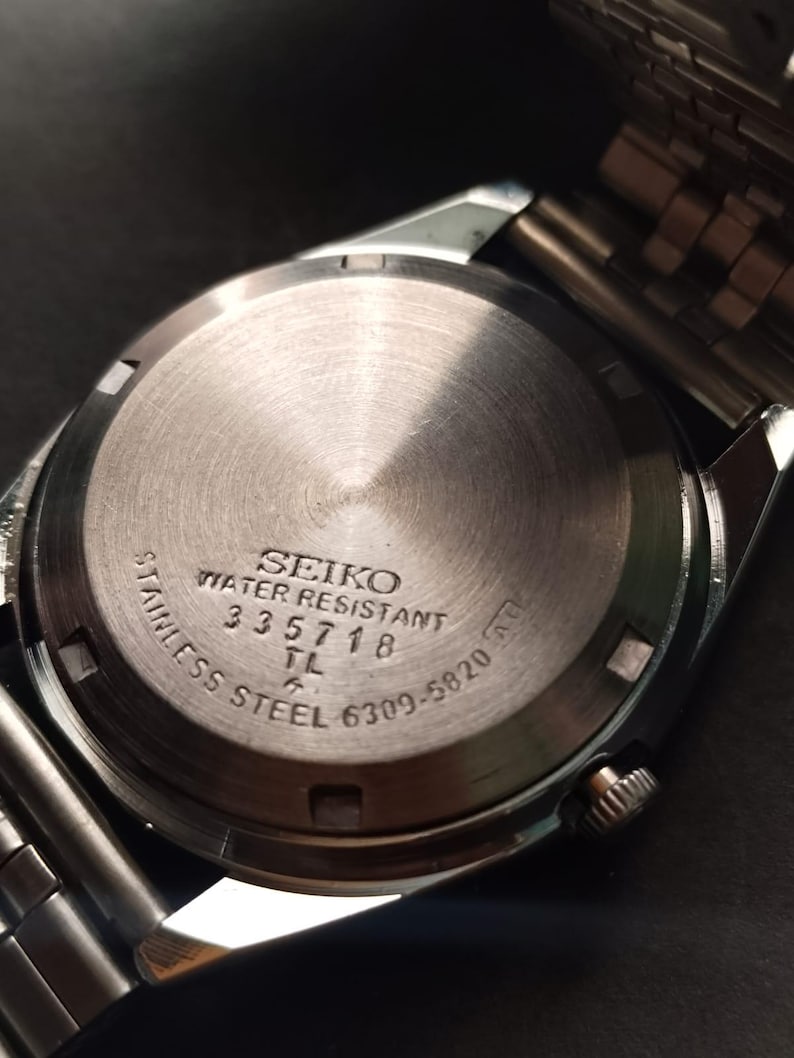 May include: Close-up of the back of a Seiko stainless steel watch. The watch face is silver with the words "SEIKO WATER RESISTANT" and other text. The watch band is silver.