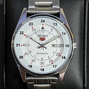 May include: A silver-toned Seiko 5 automatic wristwatch with a white face. The watch features a day and date display, a second hand, and a stainless steel band. The watch face displays the Seiko logo and Arabic numerals.