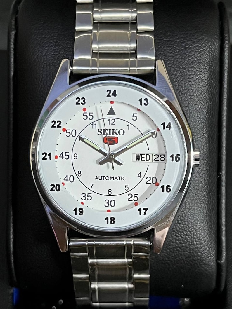 May include: A silver-toned Seiko 5 automatic wristwatch with a white face. The watch features a day and date display, a second hand, and a stainless steel band. The watch face displays the Seiko logo and Arabic numerals.