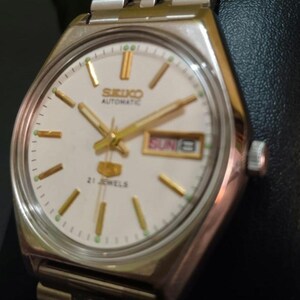 Puede incluir: Reloj de pulsera autom&aacute;tico Seiko plateado con esfera blanca. El reloj presenta agujas e indicadores dorados, una pantalla de d&iacute;a y fecha y una correa de acero inoxidable. La esfera del reloj muestra "SEIKO AUTOMATIC" y "21 JEWELS".