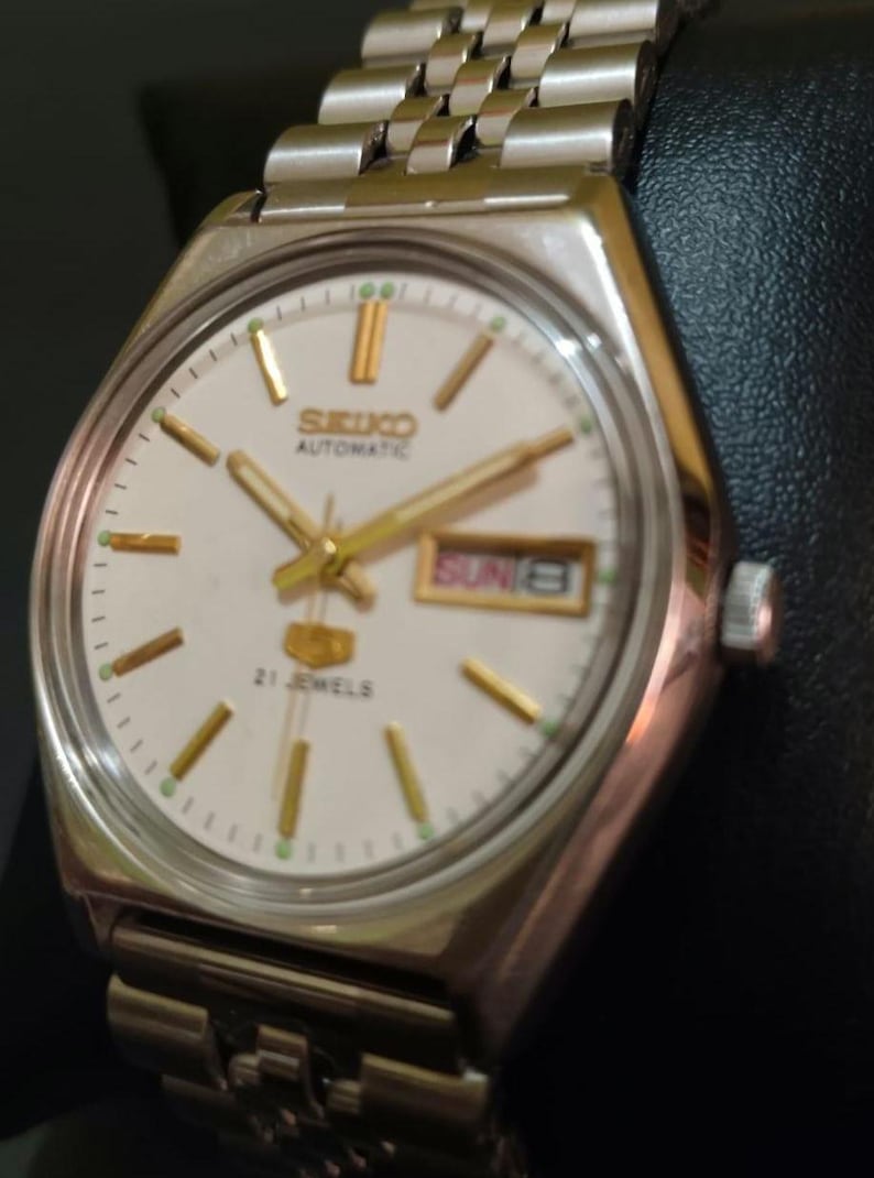 Puede incluir: Reloj de pulsera autom&aacute;tico Seiko plateado con esfera blanca. El reloj presenta agujas e indicadores dorados, una pantalla de d&iacute;a y fecha y una correa de acero inoxidable. La esfera del reloj muestra "SEIKO AUTOMATIC" y "21 JEWELS".