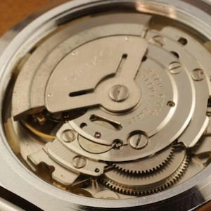 May include: The internal mechanical movement of a silver-toned Seiko watch, featuring a metallic oscillating weight, visible gears, and small screws. The text SEIKO is engraved on the rotor, with additional text indicating seventeen jewels.