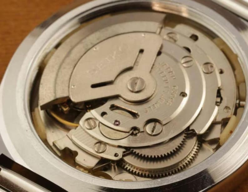 May include: The internal mechanical movement of a silver-toned Seiko watch, featuring a metallic oscillating weight, visible gears, and small screws. The text SEIKO is engraved on the rotor, with additional text indicating seventeen jewels.