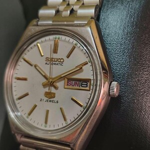 Puede incluir: Un reloj de pulsera autom&aacute;tico Seiko vintage con caja y brazalete plateados. La esfera del reloj es plateada con agujas e indicadores dorados. El d&iacute;a y la fecha se muestran a las 3 en punto. El reloj est&aacute; etiquetado como "SEIKO AUTOMATIC 21 JEWELS".