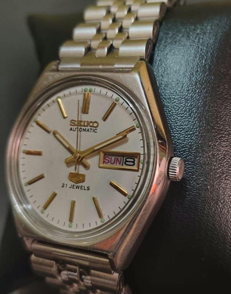 Puede incluir: Un reloj de pulsera autom&aacute;tico Seiko vintage con caja y brazalete plateados. La esfera del reloj es plateada con agujas e indicadores dorados. El d&iacute;a y la fecha se muestran a las 3 en punto. El reloj est&aacute; etiquetado como "SEIKO AUTOMATIC 21 JEWELS".