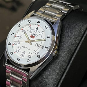 May include: A silver-toned Seiko automatic wristwatch with a white face. The watch features black numerals, a day-date display, and a silver link band. The watch face displays the brand name and the word "AUTOMATIC".