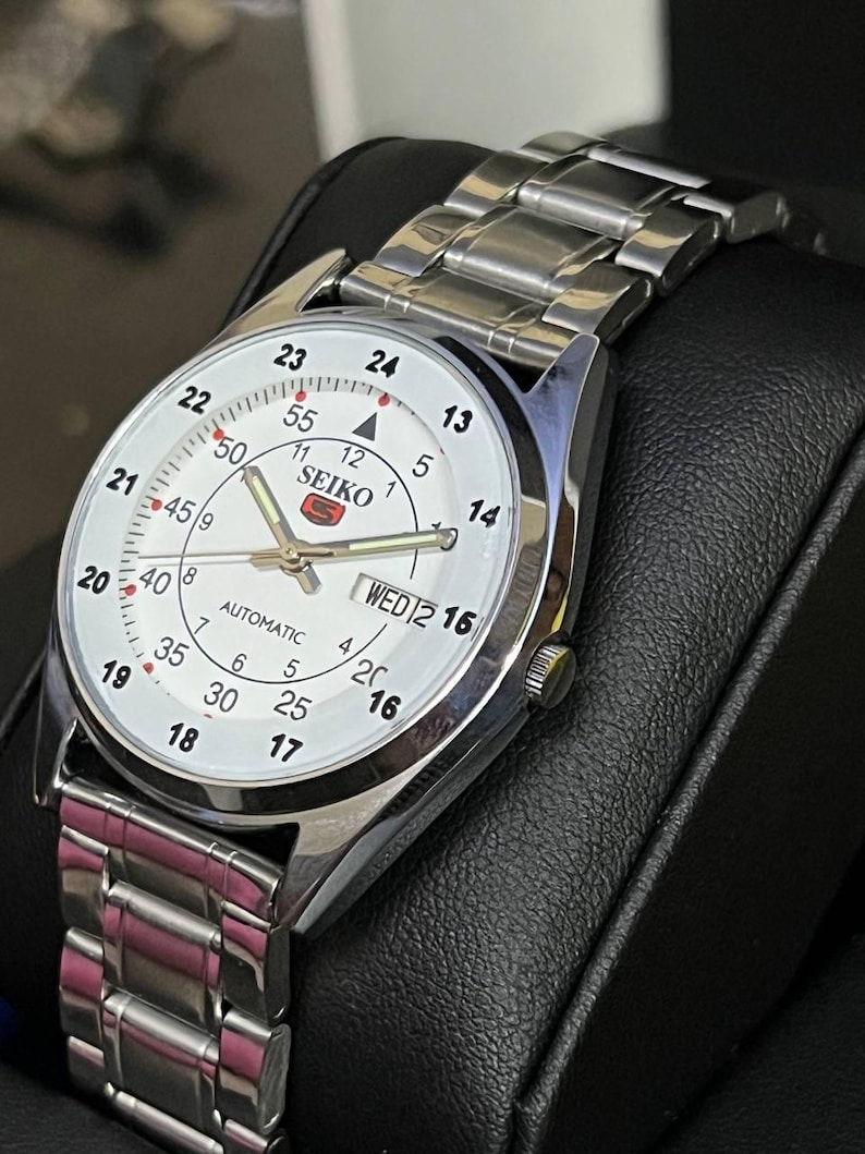 May include: A silver-toned Seiko automatic wristwatch with a white face. The watch features black numerals, a day-date display, and a silver link band. The watch face displays the brand name and the word "AUTOMATIC".