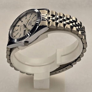 Peut inclure: Une montre-bracelet automatique Seiko argentée dotée d'un cadran crème avec des index bâtons, une lunette cannelée et un bracelet en métal à maillons multiples de style jubilé. Le cadran affiche le logo Seiko, le mot Automatic et un guichet jour-date.