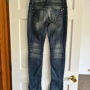 May include: Dark wash denim jeans with a faded effect, featuring decorative stitching on the back pockets and knee panels. The jeans are hanging on a white door, showcasing the back view of the garment.