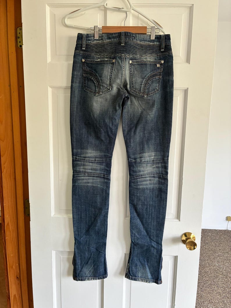 May include: Dark wash denim jeans with a faded effect, featuring decorative stitching on the back pockets and knee panels. The jeans are hanging on a white door, showcasing the back view of the garment.