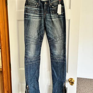 May include: Blue denim jeans with a faded wash and ribbed detailing on the front pockets and knees. The jeans have zippers at the ankles and are hanging on a white door.