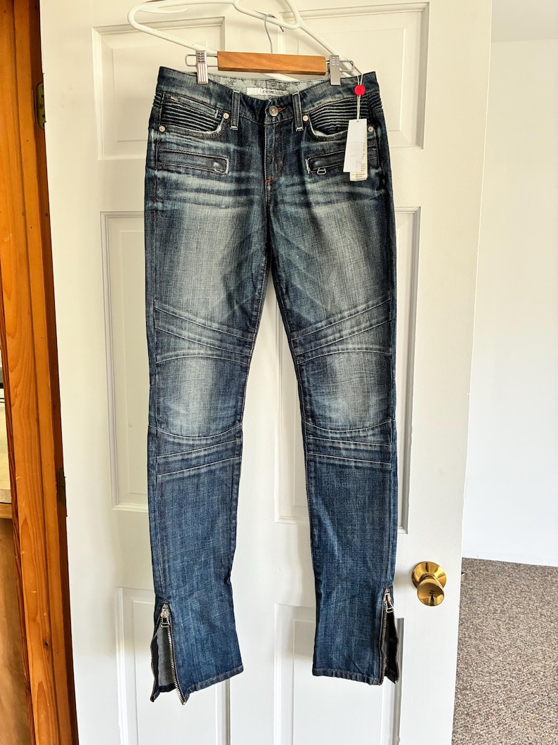 May include: Blue denim jeans with a faded wash and ribbed detailing on the front pockets and knees. The jeans have zippers at the ankles and are hanging on a white door.