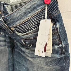 May include: Close-up of blue denim jeans with a distressed look. The jeans have a textured design on the upper thigh area and a white tag attached with a red seal. The jeans are hanging on a wooden hanger.