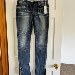 May include: Dark wash denim jeans with a distressed look, featuring ribbed detailing on the upper thighs and zippered ankles. The jeans are hanging on a white hanger against a white door.