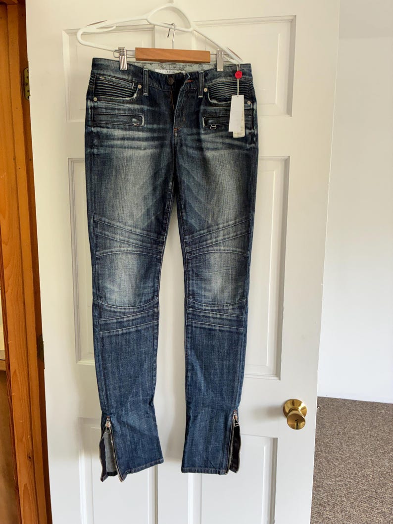 May include: Dark wash denim jeans with a distressed look, featuring ribbed detailing on the upper thighs and zippered ankles. The jeans are hanging on a white hanger against a white door.