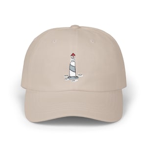 May include: A beige baseball cap featuring an embroidered lighthouse design. The lighthouse is white and gray with a red roof, set against a backdrop of blue waves. The cap has a curved brim and an adjustable strap.