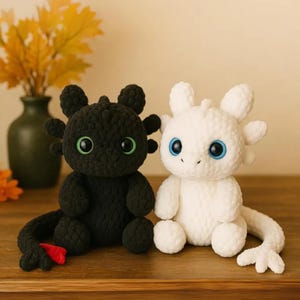 May include: Two crocheted dragon plush toys. One is black with green eyes and a red fin, the other is white with blue eyes. Both have rounded features and are sitting on a wooden surface.
