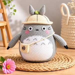 May include: A handmade crochet Totoro plushie, featuring a gray body with a white belly, pink cheeks, and a yellow hat. The plushie has black eyes and a small beige bag. It is sitting on a woven mat.