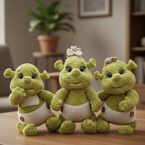Może przedstawiać: Trzy ręcznie robione zielone lalki na szydełku, przypominające popularną postać z kreskówek, są wystawione na drewnianej powierzchni. Każda lalka ma białą pieluchę i uśmiechniętą twarz. Jedna lalka ma małą kokardkę na głowie.