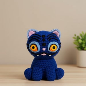 May include: A handmade blue crochet cat with large orange eyes, black markings, and a pink nose. The cat has a whimsical design and is sitting upright. The ears are light blue with pink inner details. The cat is a unique, handcrafted item.