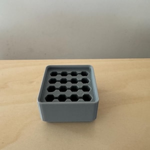 May include: A grey, rectangular organiser featuring a grid of hexagonal holes. The organiser is constructed from a solid material and is designed to hold small items. The design is simple and functional, suitable for various organisational needs.