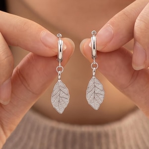 May include: A pair of silver-toned hoop earrings featuring dangling leaf-shaped charms. Each charm has a textured, matte finish with visible vein details, suspended from a small, polished circular hoop closure made of metal.