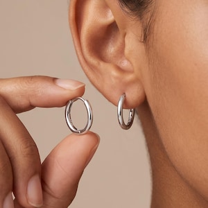 Peut inclure: Une paire de petites créoles en argent poli avec un profil lisse et arrondi. Une boucle est tenue entre les doigts, tandis que l'autre est portée au lobe de l'oreille. Le métal présente une finition brillante et un design circulaire.