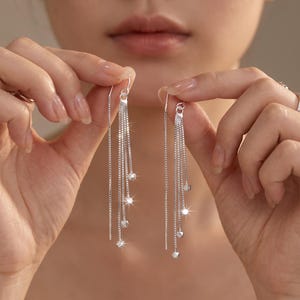 May include: A pair of silver-toned threader earrings featuring multiple delicate box chains of varying lengths. Each chain terminates in a small, faceted metallic bead, creating a cascading effect with a polished, reflective finish.