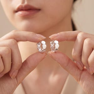 Op de afbeelding: Een paar zilveren oorringen met een modern design. Elke oorring heeft een reeks cirkelvormige, witte accenten. De oorbellen worden vastgehouden door de vingers van een persoon, waardoor hun grootte en vorm worden getoond. Een stijlvol accessoire.