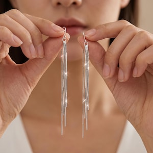 May include: Silver-toned dangle earrings with multiple fine chains cascading downwards. The earrings are held up by a person's hands, showcasing their delicate design and reflective surface. The earrings are a stylish accessory.