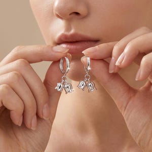 May include: Silver hoop earrings with a floral design. Each earring features a small hoop with a cluster of three tulip-shaped charms. The earrings are held up against a neutral background, showcasing their delicate details and shine.