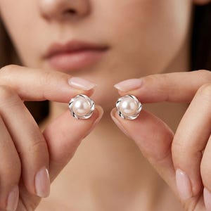 Può includere: Due orecchini a lobo in argento con un centro di perla e un design a spirale. Gli orecchini sono tenuti vicino alla fotocamera, mostrando i loro dettagli intricati e lo stile elegante. Gli orecchini sono un classico gioiello.